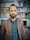 A man with a beard is standing indoors, wearing a brown blazer over a dark shirt. Behind him are shelves stocked with various items, possibly electronics or household goods, displayed in a retail setting. The background is slightly blurred, focusing attention on the man.