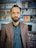 A man with a beard is standing indoors, wearing a brown blazer over a dark shirt. Behind him are shelves stocked with various items, possibly electronics or household goods, displayed in a retail setting. The background is slightly blurred, focusing attention on the man.