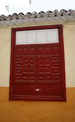 A red wooden window with decorative panels set into a wall painted in yellow and beige with a tiled roof above.