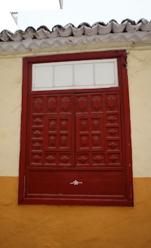 A red wooden window with decorative panels set into a wall painted in yellow and beige with a tiled roof above.