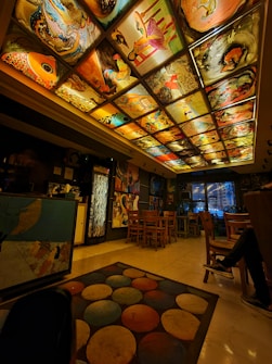 A warmly lit room with an artistic ceiling comprised of various vibrant paintings, including abstract and representational art pieces. The room features wooden chairs and tables, a colorful circular-patterned rug on a glossy floor, and eclectic wall decorations. The space has a cozy and creative atmosphere.