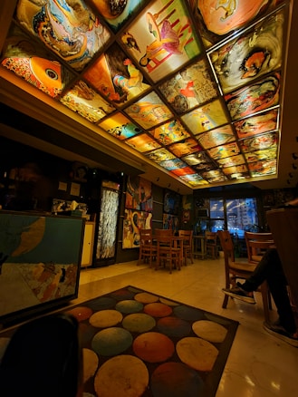 A warmly lit room with an artistic ceiling comprised of various vibrant paintings, including abstract and representational art pieces. The room features wooden chairs and tables, a colorful circular-patterned rug on a glossy floor, and eclectic wall decorations. The space has a cozy and creative atmosphere.