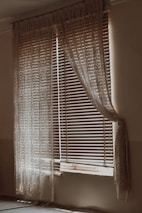 A dimly lit room with a window covered by wooden blinds and sheer lace curtains. The blinds are partially open, allowing some light to filter through, creating a gentle contrast of light and shadow on the walls and floor. The curtains drape elegantly on either side of the window, adding a vintage or rustic feel to the scene.