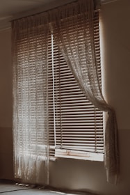 A dimly lit room with a window covered by wooden blinds and sheer lace curtains. The blinds are partially open, allowing some light to filter through, creating a gentle contrast of light and shadow on the walls and floor. The curtains drape elegantly on either side of the window, adding a vintage or rustic feel to the scene.