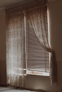 A dimly lit room with a window covered by wooden blinds and sheer lace curtains. The blinds are partially open, allowing some light to filter through, creating a gentle contrast of light and shadow on the walls and floor. The curtains drape elegantly on either side of the window, adding a vintage or rustic feel to the scene.