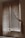A dimly lit room with a window covered by wooden blinds and sheer lace curtains. The blinds are partially open, allowing some light to filter through, creating a gentle contrast of light and shadow on the walls and floor. The curtains drape elegantly on either side of the window, adding a vintage or rustic feel to the scene.