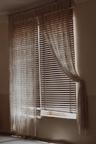 A dimly lit room with a window covered by wooden blinds and sheer lace curtains. The blinds are partially open, allowing some light to filter through, creating a gentle contrast of light and shadow on the walls and floor. The curtains drape elegantly on either side of the window, adding a vintage or rustic feel to the scene.