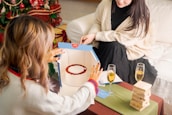 Two people are sitting in a cozy setting with festive decorations. One person is engaged with a baking activity using an instructional book and baking kit. There is a small stack of Jenga blocks on a table, alongside two glasses of champagne.