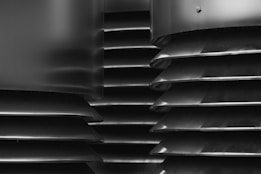 A close-up, monochromatic view of industrial metal vents or louvers with a series of horizontal slats stacked vertically. The image features smooth metallic surfaces with a slightly matte finish and a rounded bolt visible in the upper right corner, adding a mechanical element.