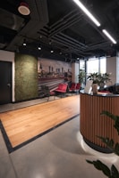 A modern office reception area featuring a sleek design with wooden accents. There is a curved wooden reception desk in the foreground and a set of red and gray chairs lined up against the wall. A green wall with plants provides a natural touch, and a large photograph of a cityscape adds a decorative element. The ceiling reveals industrial-style piping and linear lighting, while a large window allows natural light to enter, brightening the space.