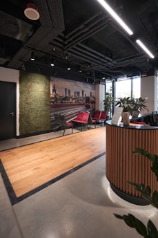 A modern office reception area featuring a sleek design with wooden accents. There is a curved wooden reception desk in the foreground and a set of red and gray chairs lined up against the wall. A green wall with plants provides a natural touch, and a large photograph of a cityscape adds a decorative element. The ceiling reveals industrial-style piping and linear lighting, while a large window allows natural light to enter, brightening the space.