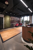 A modern office reception area featuring a sleek design with wooden accents. There is a curved wooden reception desk in the foreground and a set of red and gray chairs lined up against the wall. A green wall with plants provides a natural touch, and a large photograph of a cityscape adds a decorative element. The ceiling reveals industrial-style piping and linear lighting, while a large window allows natural light to enter, brightening the space.