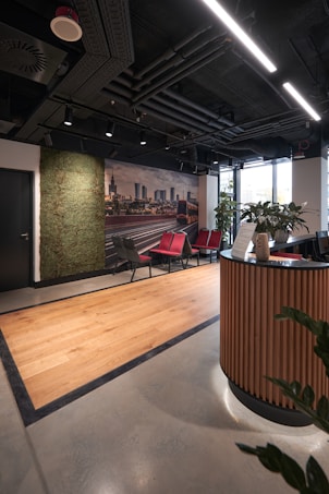 A modern office reception area featuring a sleek design with wooden accents. There is a curved wooden reception desk in the foreground and a set of red and gray chairs lined up against the wall. A green wall with plants provides a natural touch, and a large photograph of a cityscape adds a decorative element. The ceiling reveals industrial-style piping and linear lighting, while a large window allows natural light to enter, brightening the space.
