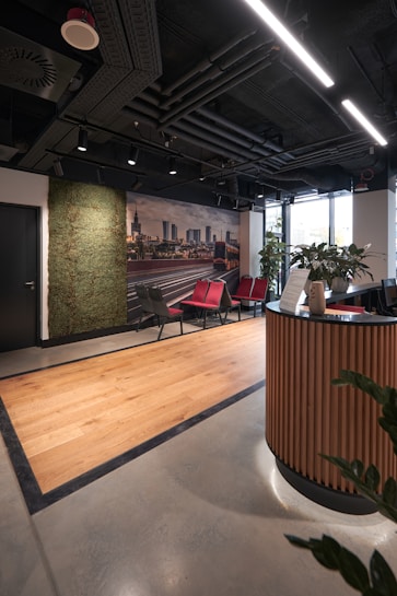 A modern office reception area featuring a sleek design with wooden accents. There is a curved wooden reception desk in the foreground and a set of red and gray chairs lined up against the wall. A green wall with plants provides a natural touch, and a large photograph of a cityscape adds a decorative element. The ceiling reveals industrial-style piping and linear lighting, while a large window allows natural light to enter, brightening the space.
