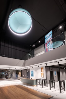 A modern interior space featuring a large, circular, teal-colored ceiling light with a glowing outer rim. Below is a spacious area with a wooden floor, a reception desk with a ribbed front, and turnstiles. The walls are grey, complemented by a textured tile floor. There are posters and signs affixed to the walls and a large glass entrance. The overall design is sleek and contemporary, with a minimalistic and open layout.