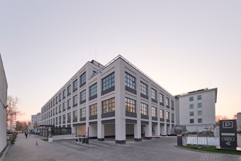 A modern, multi-story building with large grid-like windows, surrounded by a paved plaza area. The architecture is clean with a white and gray color scheme. A few people are walking along the sidewalk, and there are parking signs and structures indicating a parking area.