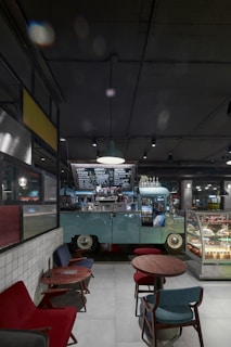 A cozy indoor cafe setting with a vintage van repurposed as a coffee counter. The van is mint green, parked beneath modern industrial lighting, with a handwritten menu visible above. In the foreground, there are small round wooden tables with colorful upholstered chairs, and a display case filled with assorted desserts.