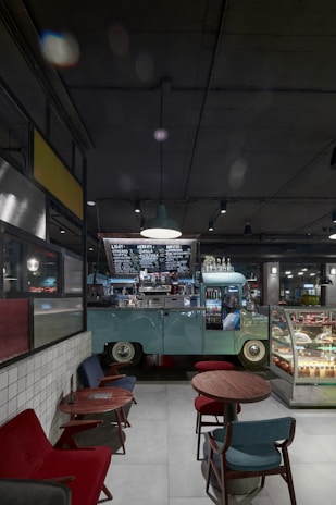 A cozy indoor cafe setting with a vintage van repurposed as a coffee counter. The van is mint green, parked beneath modern industrial lighting, with a handwritten menu visible above. In the foreground, there are small round wooden tables with colorful upholstered chairs, and a display case filled with assorted desserts.