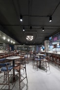 A modern restaurant interior featuring multiple wooden tables and chairs, a sleek bar area with stools, and contemporary hanging lights. The ceiling is industrial with exposed ductwork, and the space is well-lit with a spacious, open layout.