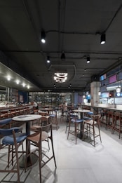 A modern restaurant interior featuring multiple wooden tables and chairs, a sleek bar area with stools, and contemporary hanging lights. The ceiling is industrial with exposed ductwork, and the space is well-lit with a spacious, open layout.