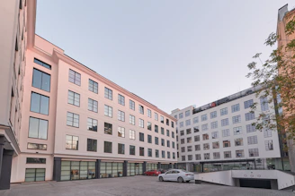 A modern multi-story building with large reflective windows and a light, neutral facade surrounds a courtyard area. There are a few cars parked on the paved courtyard, and some greenery is visible along the edge. The architecture is clean and minimalist, suggesting a business or residential function.