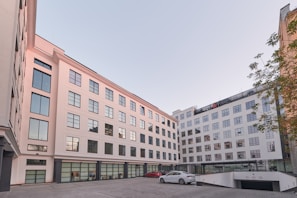 A modern multi-story building with large reflective windows and a light, neutral facade surrounds a courtyard area. There are a few cars parked on the paved courtyard, and some greenery is visible along the edge. The architecture is clean and minimalist, suggesting a business or residential function.