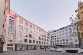 A modern multi-story building with large reflective windows and a light, neutral facade surrounds a courtyard area. There are a few cars parked on the paved courtyard, and some greenery is visible along the edge. The architecture is clean and minimalist, suggesting a business or residential function.
