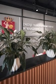 A modern reception area featuring a wooden counter with a black top. Two large potted plants in white geometric planters are prominently displayed on the counter. The background includes a wall with a metal mesh design and signage in Polish language, along with a decorative emblem featuring a crowned figure.