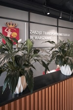 A modern reception area featuring a wooden counter with a black top. Two large potted plants in white geometric planters are prominently displayed on the counter. The background includes a wall with a metal mesh design and signage in Polish language, along with a decorative emblem featuring a crowned figure.