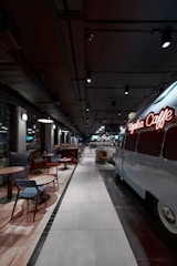 A modern, stylish cafe with an industrial design features a retro van converted into a seating or serving area. Comfortable chairs and small tables line one side, while pendant lights and exposed ceilings create an urban vibe.