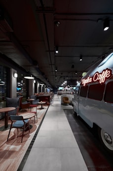 A modern, stylish cafe with an industrial design features a retro van converted into a seating or serving area. Comfortable chairs and small tables line one side, while pendant lights and exposed ceilings create an urban vibe.