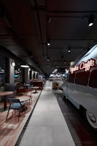 A modern, stylish cafe with an industrial design features a retro van converted into a seating or serving area. Comfortable chairs and small tables line one side, while pendant lights and exposed ceilings create an urban vibe.