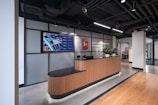 A modern office reception area features a sleek wooden reception desk topped with several small potted plants. Above the desk, a large digital screen displays a map with labeled areas. The backdrop consists of a wall with industrial-style metallic paneling and an emblem. Overhead lights illuminate the space, highlighting the wood-paneled flooring and the modern, exposed ceiling with visible ductwork.