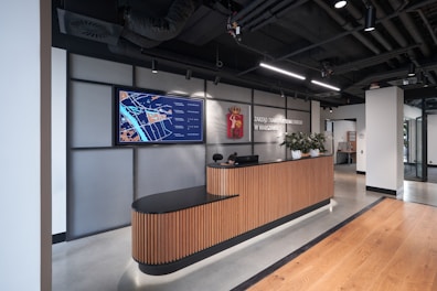 A modern office reception area features a sleek wooden reception desk topped with several small potted plants. Above the desk, a large digital screen displays a map with labeled areas. The backdrop consists of a wall with industrial-style metallic paneling and an emblem. Overhead lights illuminate the space, highlighting the wood-paneled flooring and the modern, exposed ceiling with visible ductwork.