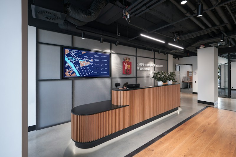 A modern office reception area features a sleek wooden reception desk topped with several small potted plants. Above the desk, a large digital screen displays a map with labeled areas. The backdrop consists of a wall with industrial-style metallic paneling and an emblem. Overhead lights illuminate the space, highlighting the wood-paneled flooring and the modern, exposed ceiling with visible ductwork.