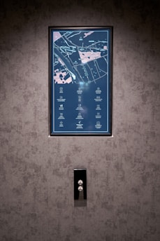 A vertical display screen is mounted on a textured grey wall, showing a detailed map with icons and text in a combination of light blue and pink colors. Below the screen, there is a sleek, reflective panel with two buttons, possibly serving as elevator controls.