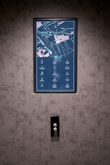 A vertical display screen is mounted on a textured grey wall, showing a detailed map with icons and text in a combination of light blue and pink colors. Below the screen, there is a sleek, reflective panel with two buttons, possibly serving as elevator controls.