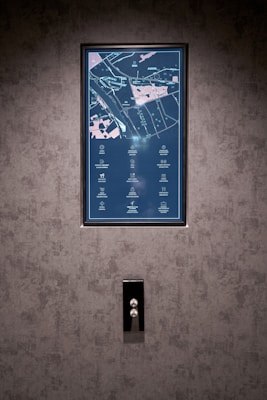 A vertical display screen is mounted on a textured grey wall, showing a detailed map with icons and text in a combination of light blue and pink colors. Below the screen, there is a sleek, reflective panel with two buttons, possibly serving as elevator controls.