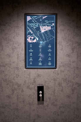 A vertical display screen is mounted on a textured grey wall, showing a detailed map with icons and text in a combination of light blue and pink colors. Below the screen, there is a sleek, reflective panel with two buttons, possibly serving as elevator controls.
