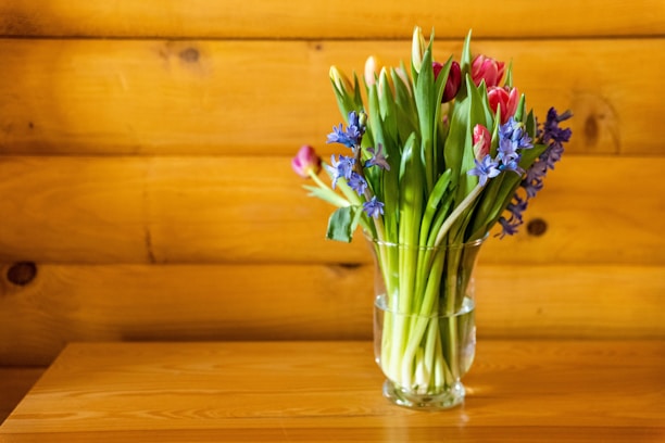 A sleek transparent glass vase filled with fresh spring flowers on a wooden table.