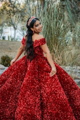 A woman with long dark hair is wearing an elegant, sparkly red gown. She is adorned with a matching red tiara and earrings. Her pose is joyful, with a bright smile. The background features tall grasses and some out-of-focus trees, suggesting a natural setting.