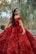 A woman with long dark hair is wearing an elegant, sparkly red gown. She is adorned with a matching red tiara and earrings. Her pose is joyful, with a bright smile. The background features tall grasses and some out-of-focus trees, suggesting a natural setting.