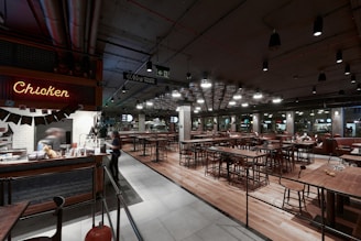 A spacious, contemporary restaurant interior with high ceilings and industrial-style lighting. A neon sign reading 'Chicken' is visible on the left above a counter where a person is serving food. Several wooden tables and chairs fill the space, with a few patrons seated along the right side. The decor is modern with a mix of metal and wood elements, and the walls have large glass windows.