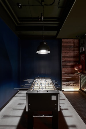 A foosball table stands in the center of a dimly lit room. The lighting fixture hanging above casts a focused beam onto the table, creating a dramatic spotlight effect. The walls are painted dark blue, contrasting with the wooden panels and shelves on the right side, where some decorative objects, such as two globe-like items, are placed. The ceiling features exposed pipes and ducts, contributing to an industrial aesthetic.
