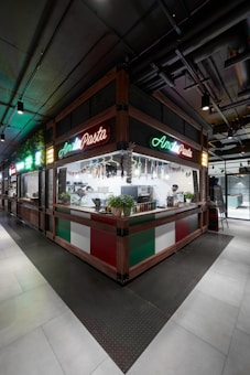 A corner kiosk with vibrant neon signs displaying the words 'Amal Pasta' is seen. The counter features Italian flag colors, and there are pots of fresh herbs placed on it. Hanging dried meats decorate the area while chefs work in the open kitchen behind the counter.
