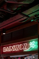Neon sign with the word 'BARSZCZ' illuminated in front of a red and green background with a decorative leaf design. The setting appears to be indoors, showcasing metal ceiling infrastructure.