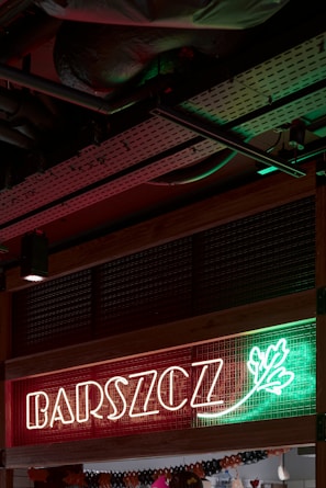 Neon sign with the word 'BARSZCZ' illuminated in front of a red and green background with a decorative leaf design. The setting appears to be indoors, showcasing metal ceiling infrastructure.