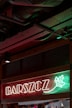 Neon sign with the word 'BARSZCZ' illuminated in front of a red and green background with a decorative leaf design. The setting appears to be indoors, showcasing metal ceiling infrastructure.