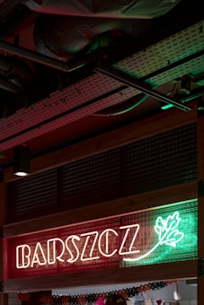 Neon sign with the word 'BARSZCZ' illuminated in front of a red and green background with a decorative leaf design. The setting appears to be indoors, showcasing metal ceiling infrastructure.