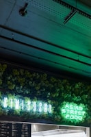 A neon sign displaying 'KIMI SUSHI' and 'ASIAN STREET FOOD' is illuminated against a backdrop of lush green foliage. The dim setting creates a contrast, highlighting the vibrant greens from the sign. The ceiling above features exposed industrial elements.